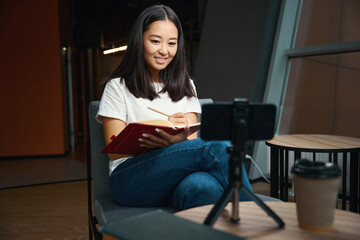 Happy stylish female is taking notes in notebook while studying online