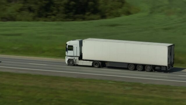 One Semi Truck with white trailer and cab driving, traveling alone on asphalt straight empty road through plain field, highway top down view follow vehicle aerial footage. Freeway trucks traffic