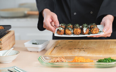 Selective focus on japanese food called takoyaki in plate from hot pan in hands of anonymous young chef man, in black uniform, showing dishes on table at kitchen restaurant