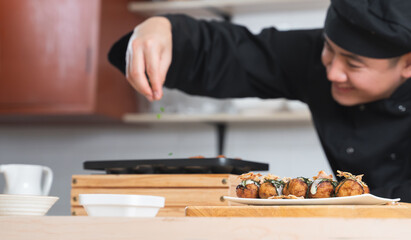 Selective focus on japanese food called takoyaki in plate from hot pan on table. Asian young chef man, in black uniform, cooking takoyaki dishes on blur background, at kitchen restaurant