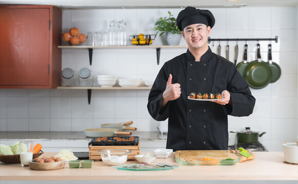 Asian Young Handsome Chef Man, In Black Uniform, Smiling Thumbs Up And Holding On Japanese Food Called Takoyaki In Plate Ready To Serve From Hot Pan With Ingredients On Table At Kitchen Restaurant