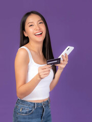 Beautiful Asian brunette woman cute girl in white tank top holding credit card and smartphone ,mobile phone uses a online banking for pay online shopping isolated on purple background