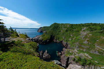 Fototapeta na wymiar A hiking trail along the ocean with waves crashing on the jagged rocks. The calm water and sky are blue with white clouds. There are trees, rocks, grass, and eroding cliff along the edge of the ocean.