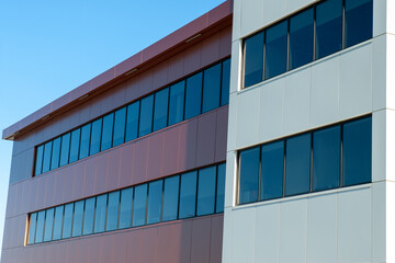 The exterior wall of a large building with tan colored panels, square glass windows with the cloudy blue sky reflecting in the windows, and rusty red composite metal panels. The trim is beige metal.