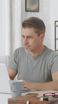 Older man sitting at desk in bright room working with laptop computer in home office. Entrepreneur managing business online.  Mature age, middle age, mid adult casual man in 50s, happy smiling.