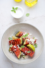Beige plate with grilled watermelon batons and grated cheese, vertical shot on a light-beige marble background, elevated view