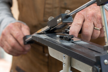 A man using a fixed angle knife sharpener kitchen tools at a farmer's market to sharpen knives for customers. The stainless steel kit has a long narrow holder for water stones of various grits. 