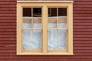 An exterior wall with red colored horizontal cape cod clapboard siding and a double hung window. The closed vintage glass window has yellow and cream colored trim and molding with a window blind.