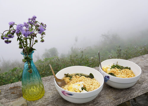 Delicious bowls of instant ramen cooked with egg and vegetables served on a wood bench in cold foggy sky of Ta Xua, Vietnam