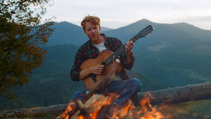 Happy guy sing camp song in mountains. Positive hipster play guitar on nature.