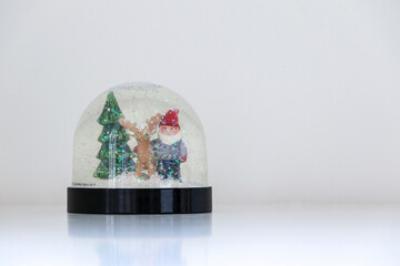 A sparkling and glittering Christmas snow globe with snow falling on Santa Claus, a reindeer, and a Christmas tree, on white snowy background, copy space on the right