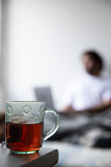 cup with tea on a table, in the background a man appears out of focus typing on his computer sitting on the bed