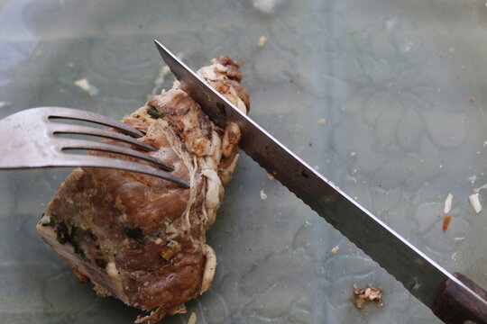 Corte con tenedor y cuchillo de una porciòn de asado de cerdo a la parrilla sobre el plato, hecho en casa en forma rùstica, muestra un original diseño culinario con fondo gris