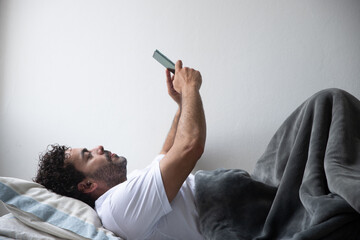 man lying on his bed holding his phone with both hands up, man working at his home lying on the bed