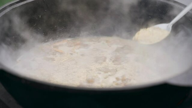 The cook sprinkles and mixes with a spoon of seasoning for pilaf, which is cooked in a cast-iron cauldron. Close-up.