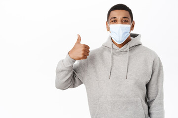 Covid awareness, healthcare concept. Image of happy african american man in medical mask, shows thumbs up