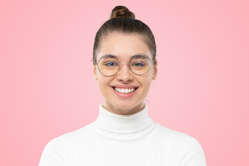 Headshot of young woman wearing glasses and white turtleneck, smiling at camera on pink background