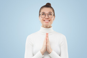 Smiling girl in eyeglasses looking above praying, holding palms together, asking to fulfill dream