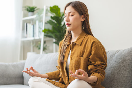 Peaceful asian young woman, girl hands in calm pose sitting practice meditating in lotus position on sofa at home, meditation, exercise for wellbeing, healthy care. Relaxation, happy leisure people.