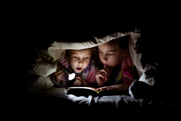 Two sister girls read a book under a blanket in the dark with a flashlight.