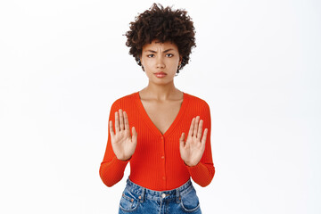 Portrait of serious Black girl shows stop gesture, disapprove smth, rejects and prohibits, stands over white background