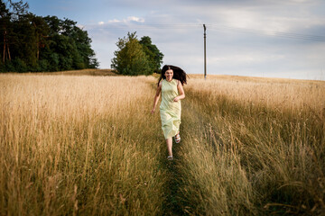 a beautiful girl with long dark hair in a light green dress runs through a field on a summer evening