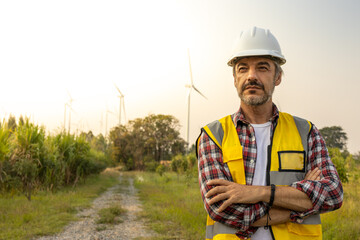 Aืn Engineer standing by the wind turbine in the renewable power farm. He is wearing a white hard...