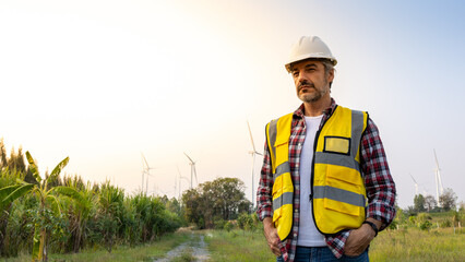 Aืn Engineer standing by the wind turbine in the renewable power farm. He is wearing a white hard hat and yellow transparent vest.