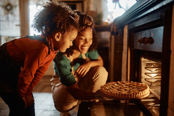 Happy black little girl and her mother taking out sweet pie from the oven in kitchen.