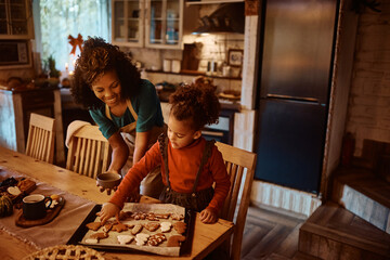 Black little girl making gingerbread cookies with her mother in kitchen.