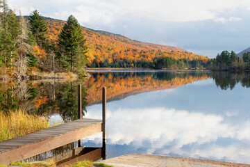 Tranquil autumn scene in White Mountain National Forest of New Hampshire. Wooden boat dock with reflection of sky and colorful fall foliage on calm surface of scenic Long Pond.