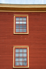 The exterior of a red colored cottage with wooden wall covered in horizontal clapboard siding. There are two vintage windows with multiple glass panes. The decorative trim is yellow and orange colored