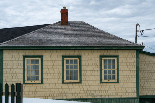 A vintage yellow cedar shake clapboard house with a grey shingled wooden hip roof. The building has a red brick chimney and multiple glass windows.  The sky in the background is blue with clouds.
