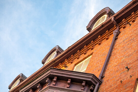 An upward view of a historic residential building with red brick, yellow and green wooden decorative style wood trim and glass single hung windows with vintage dormers. The sky is blue and cloudy.