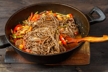 Soba with vegetables in soy sauce with spices in a pan with a spatula on a wooden stand on a wooden table.