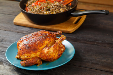 Whole baked chicken carcass stuffed with pear in soy sauce with a crispy crust on a plate next to soba in a pan with vegetables on a wooden table.