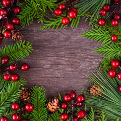 Fototapeta na wymiar Christmas frame with evergreen branches, red berries and pine cones. Top down view on a square dark wood background with copy space.