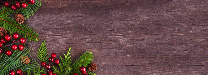 Winter corner border with evergreen branches, red berries and pine cones. Above view on a dark wood banner background with copy space. - obrazy, fototapety, plakaty