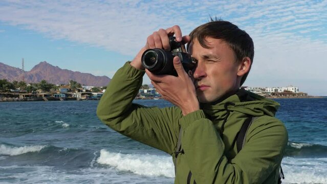 Male photographer takes pictures with camera of urban view, local sightseeing attraction on sea coast. Man tourist shooting landmarks of seaside town standing near water. Travel photos of resort city