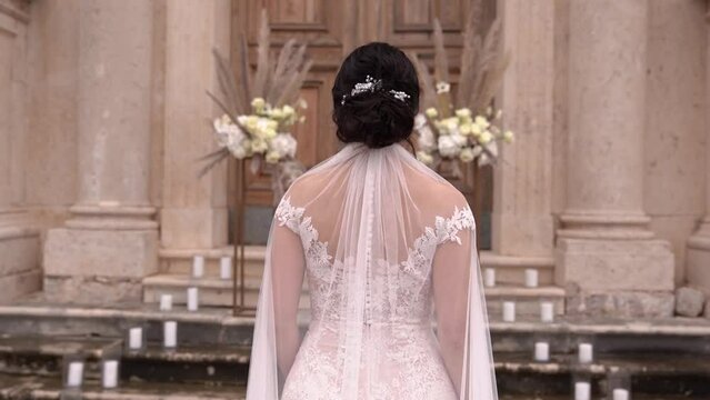 Bride in a white dress with a veil goes to the entrance of the church. Back view
