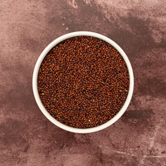 Top view of a white bowl filled with red quinoa on a maroon tabletop.