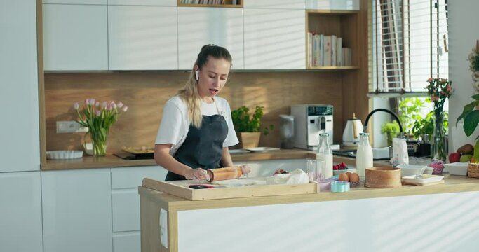 Pretty adorable young woman in modern new headphones cellphone listening to music relaxing while cooking rolling homemade dough with rolling pin singing insinging song holding beater whisk microphone.