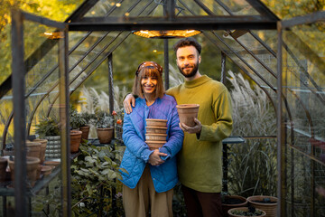 Obraz premium Portrait of a young couple stand together with clay jugs in small greenhouse for growing flowers in garden. Hobby or small family business of growing flowers concept
