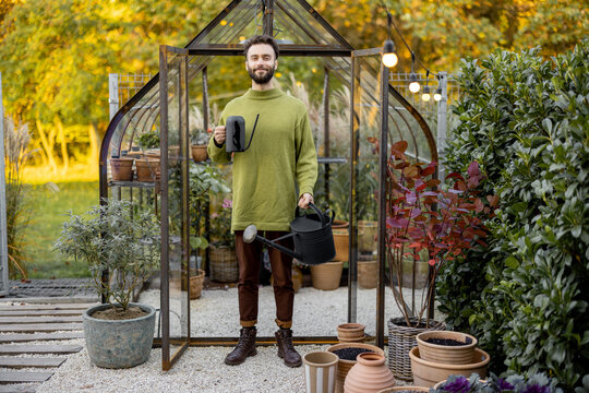 Portrait Of A Stylish Guy Stand With Watering Can In Front Of A Tiny Greenhouse For Growing Plants In Garden. Hobby, Work In Garden Concept