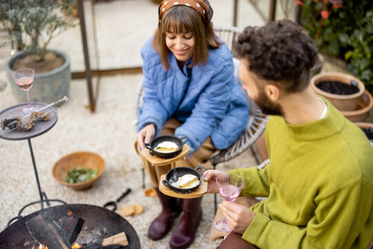 Young Couple Have A Dinner, Eating Hot Cheese With Bread And Drink Wine While Sitting Together By The Fire In Garden