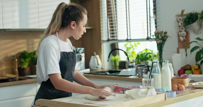 Beautiful young mother in modern kitchen at home wearing apron having fun rolling homemade dough on wooden surface using rolling pin baking cooking tasty cookies watching movie film on tablet.