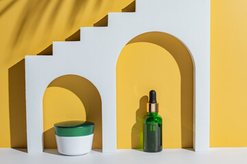 green glass bottle with a pipette and cream jar in white decorative arches on a yellow background....
