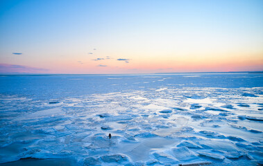 Fototapeta na wymiar Aerial view - lonely man walk on ice on sunset over a frozen sea. Winter landscape on seashore during dusk. View from above of melting ice in ocean on sunrise. Global warming. Colorful skyline.