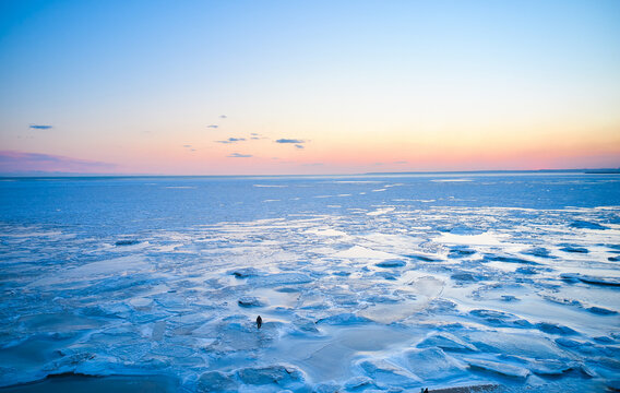 Aerial view - lonely man walk on ice on sunset over a frozen sea. Winter landscape on seashore during dusk. View from above of melting ice in ocean on sunrise. Global warming. Colorful skyline.