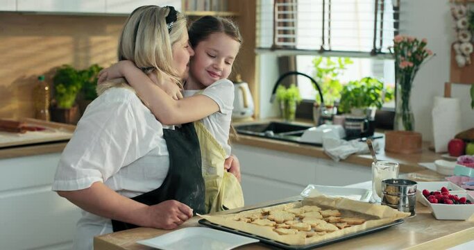 Happy kid hugging with middle aged granny in modern kitchen while taking cookies from baking tray to white beautiful plate. Curious girl tasting delicious biscuit cookies.